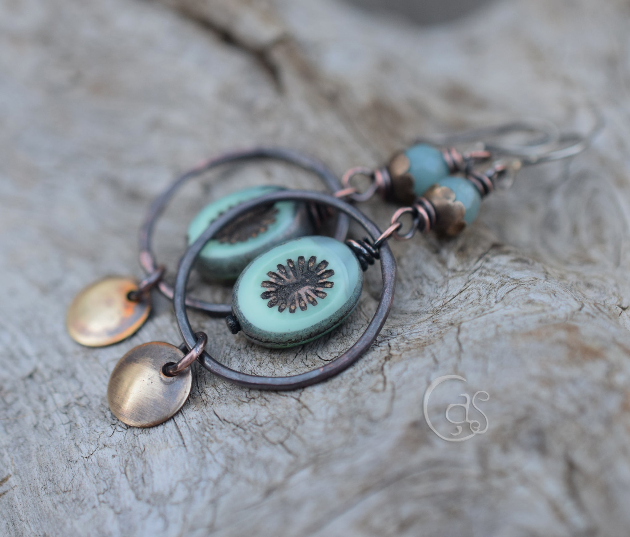 A pair of earrings featuring large copper hoops, aqua blue oval beads with floral designs, and small metal disc charms, displayed on a rustic wooden surface.