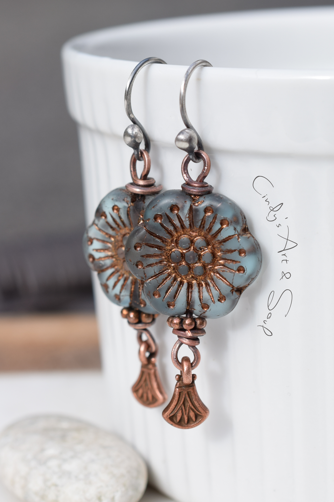 Blue and copper flower earrings with copper dangles hanging from a white cup next to a gray stone. 