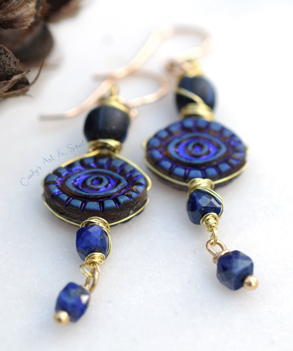 A pair of handmade dangle earrings with dark blue and purple concentric spiral beads, gold wire wrapping, and faceted blue beads, displayed on a white textured surface.