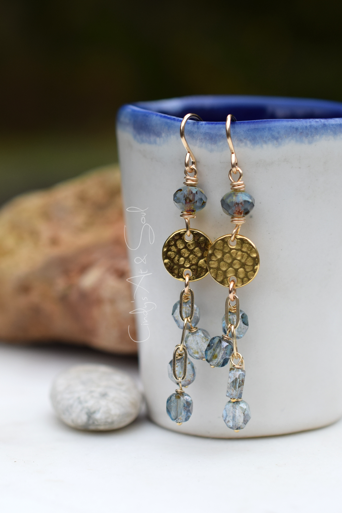 Gold and blue beaded earrings hanging from a ceramic mug with a blurred natural background.
