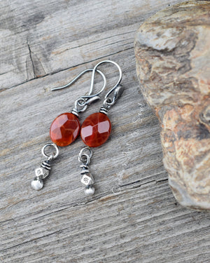 Dark orange gemstone earrings made with silver. The background is wood and there is a rock next to the earrings. 