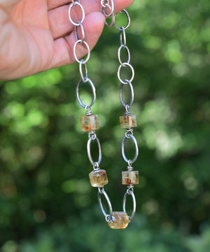 Hand holding a silver chain with amber-colored beads against a blurred green background
