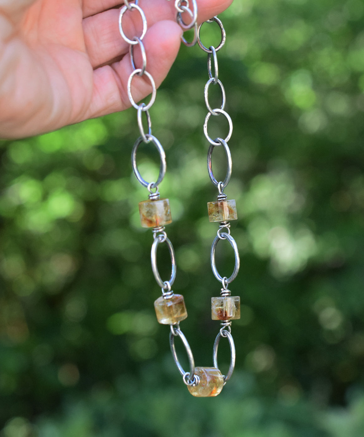Hand holding a silver chain with amber-colored beads against a blurred green background