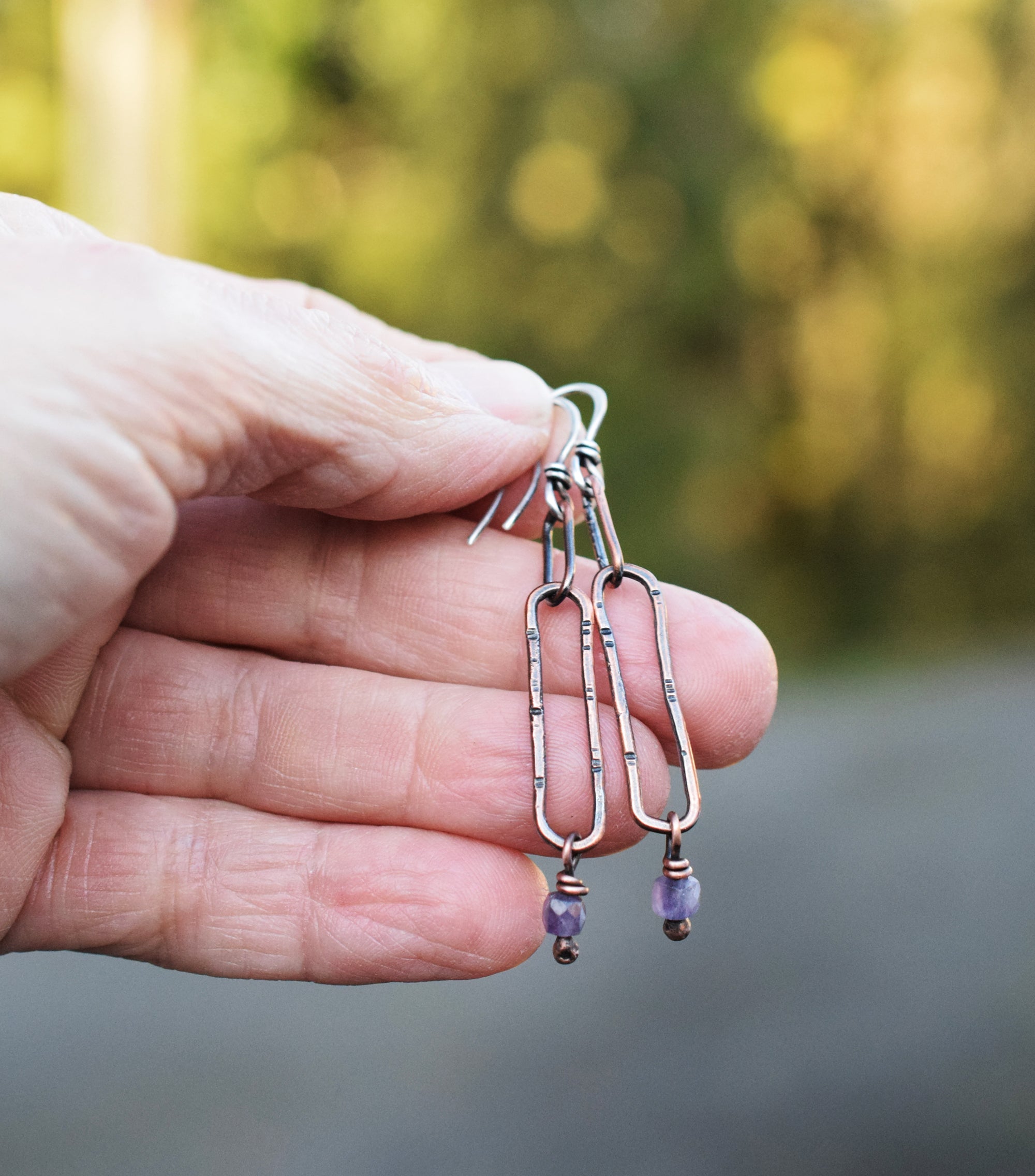 Hand holding a pair of earrings with purple beads against a blurred natural background