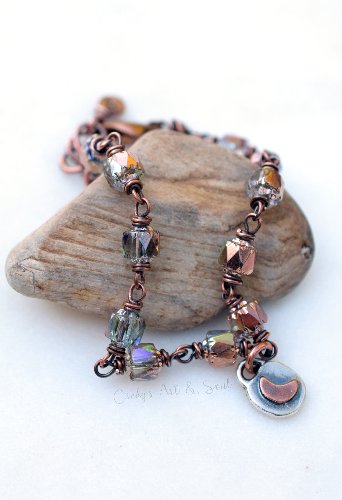 A handmade beaded bracelet with copper wire, faceted beads, iridescent glass beads, and a small silver moon charm, displayed on a natural stone against a white background.