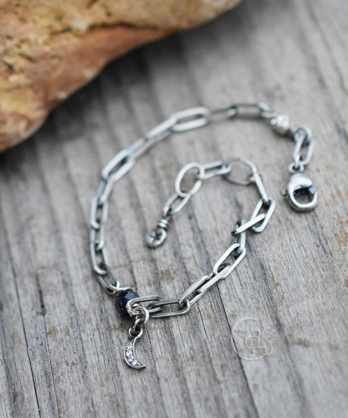 Silver bracelet with blue gemstone and crescent moon charm on a wood background next to a brown rock. 