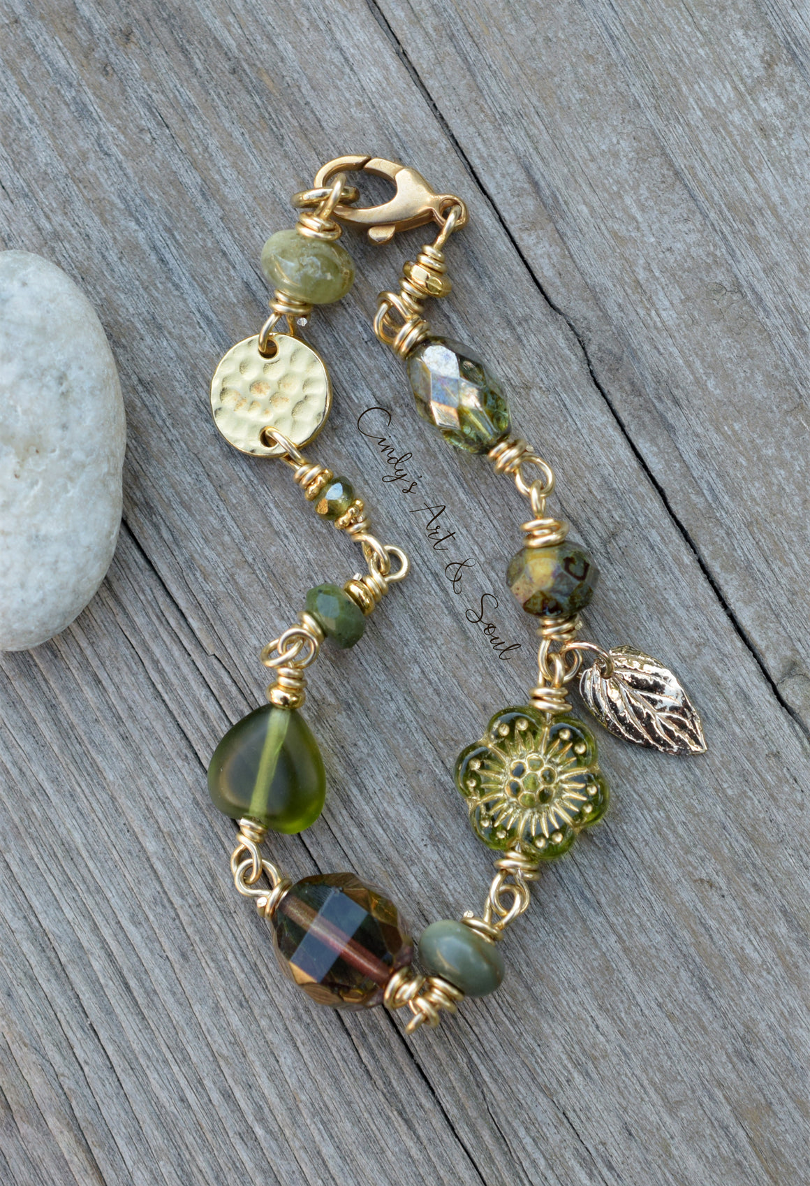 A handmade beaded bracelet with gold-toned wire wrapping, featuring green and brown beads, a hammered gold disc, a leaf charm, and a faceted bead, displayed on a weathered gray wooden surface next to a white stone.