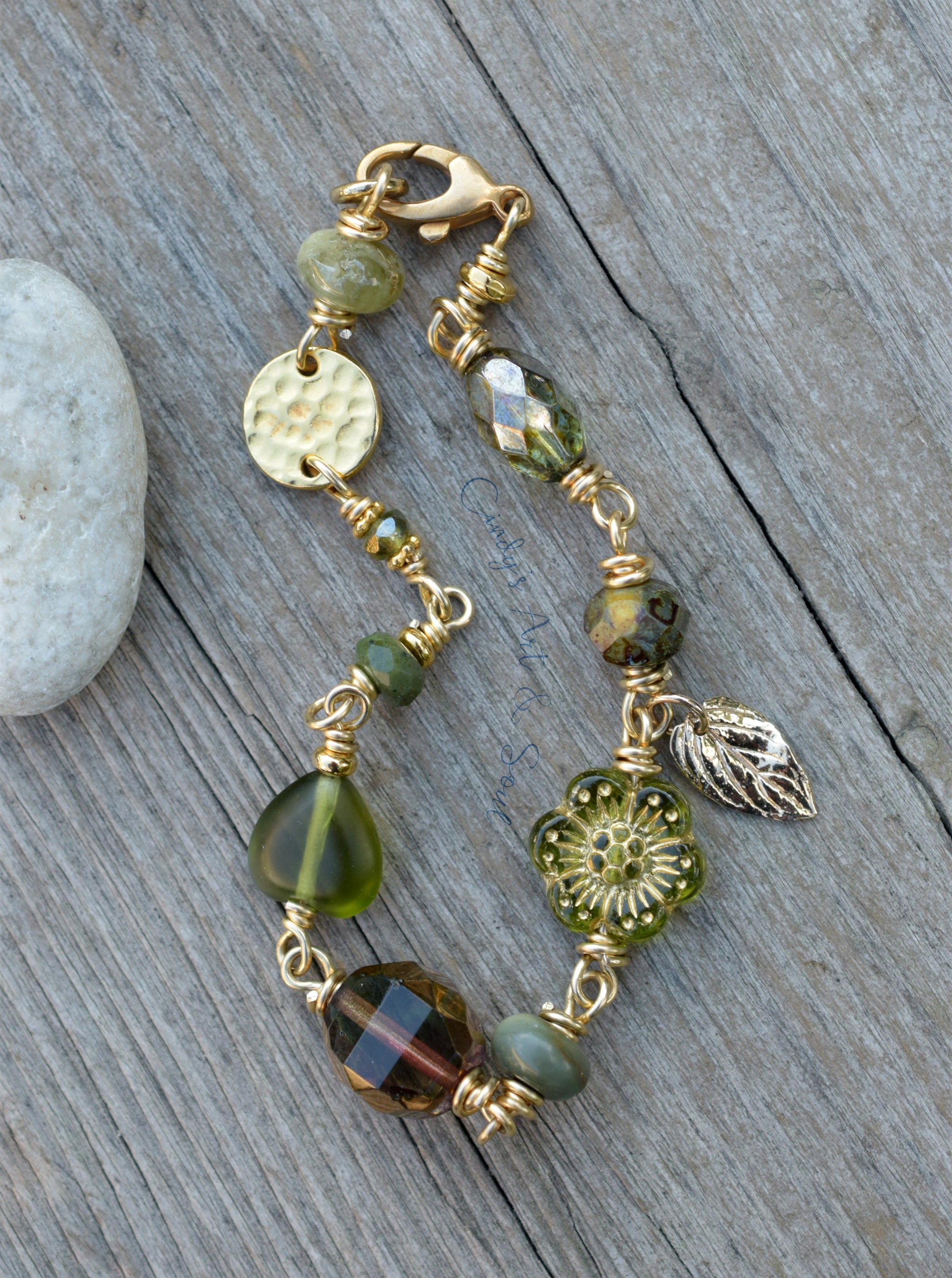 A beaded bracelet with gold-toned wire wrapping, green faceted beads, a hammered gold disc, a floral gold charm, and a silver-toned leaf charm, displayed on a weathered gray wooden surface next to a white stone.