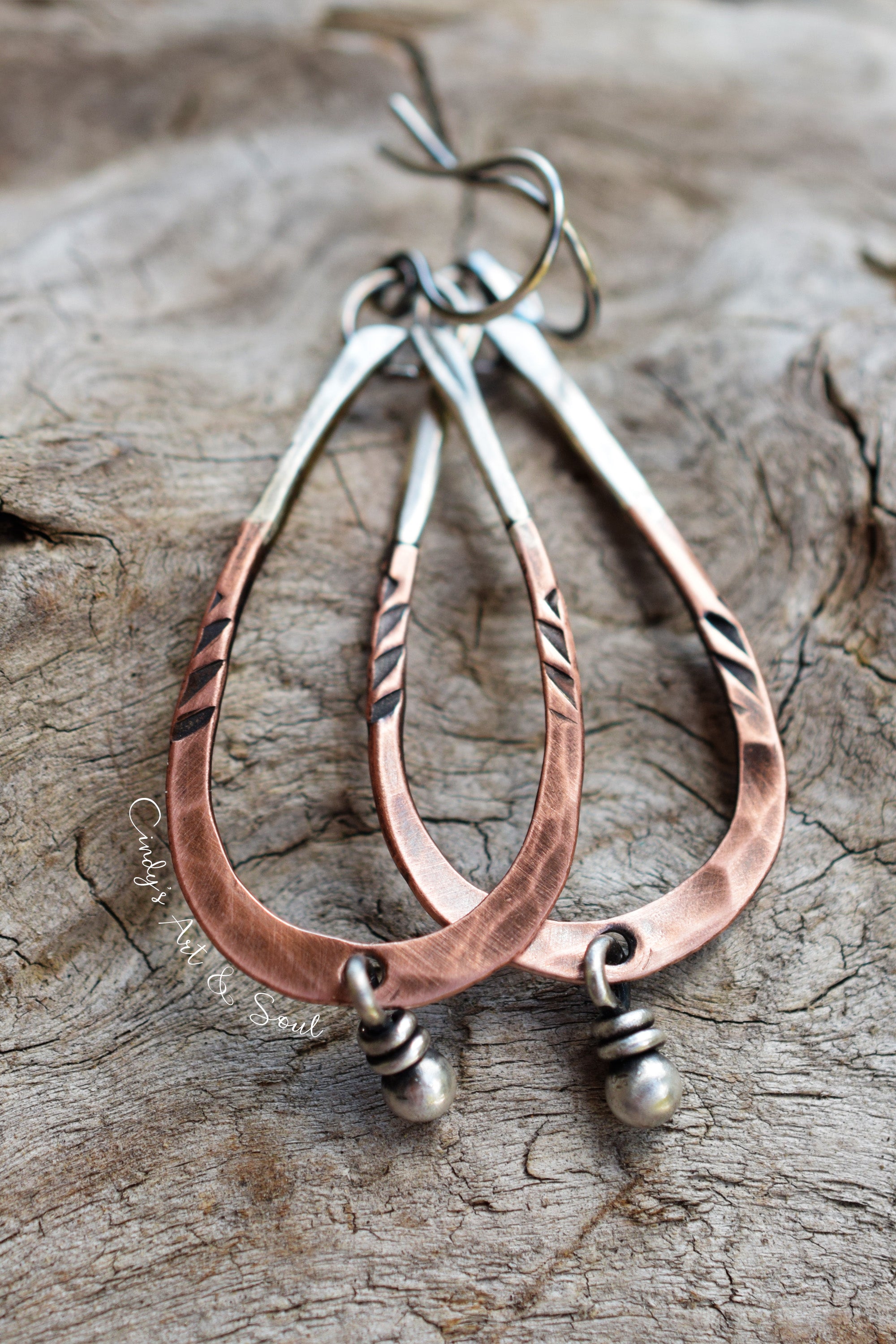 Handmade teardrop dangle earrings made of hammered copper and silver-toned metal, displayed on a textured wood background.