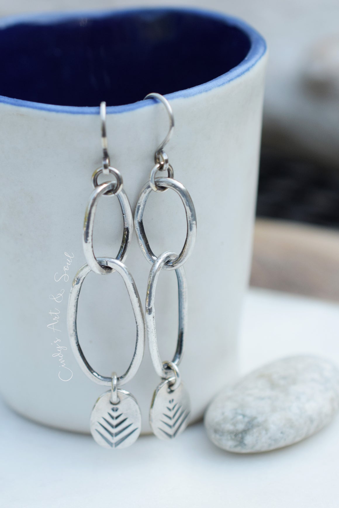 Sterling silver dangle earrings with interlocking oval links and chevron-patterned teardrop charms, hanging in front of a white ceramic mug with a blue interior.