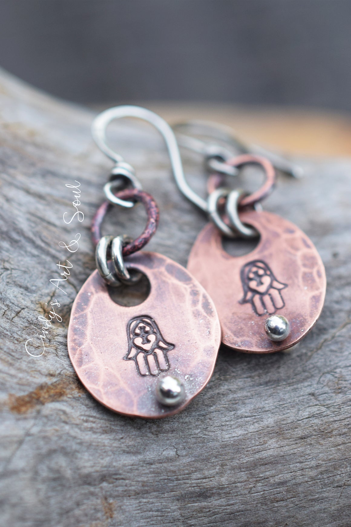 Handmade copper and silver dangle earrings with stamped Hamsa hand designs, resting on a textured gray wood surface.