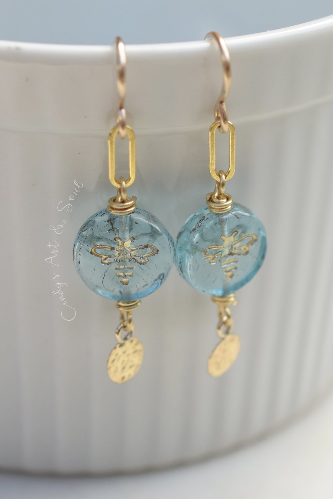 A pair of gold-tone drop earrings with round light blue beads featuring an embossed bee design, hanging from gold hooks in front of a white ribbed ceramic cup.