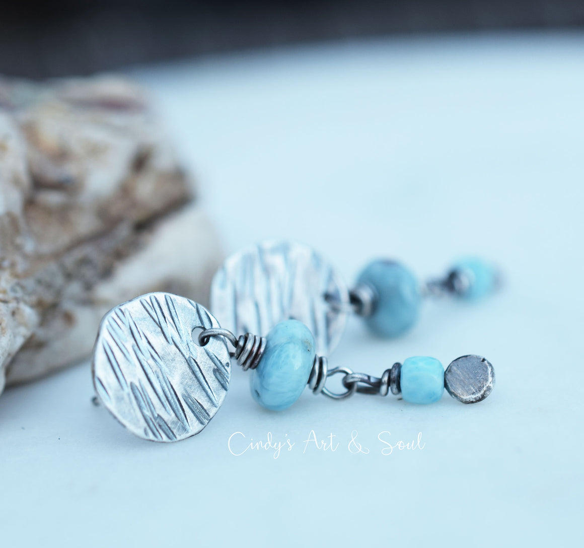 A pair of handmade dangle earrings featuring textured silver disc charms, light blue beads, and small silver spacer beads, displayed on a light blue surface with a blurred stone prop in the background.