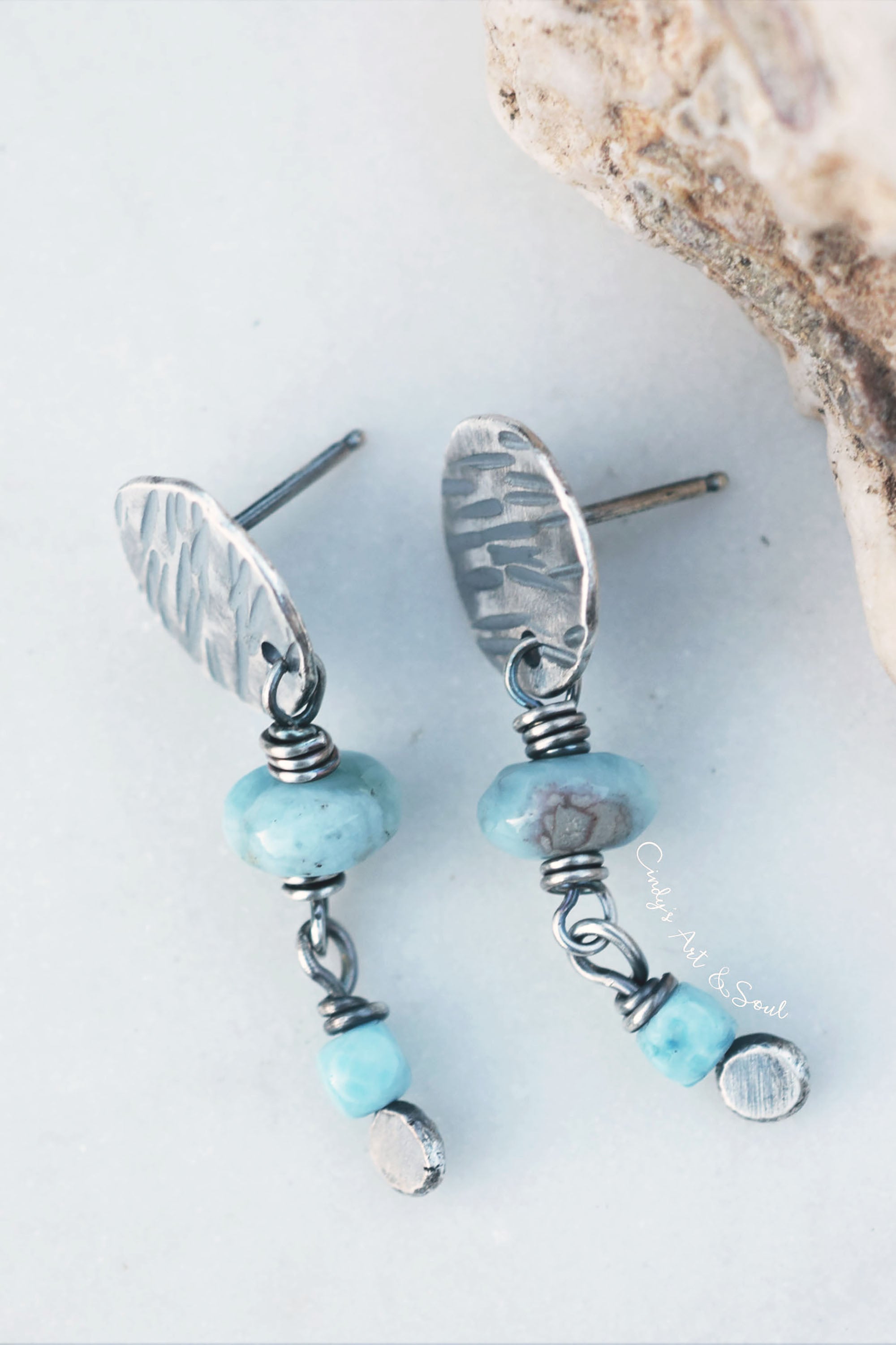 A pair of handmade dangle earrings featuring hammered silver-toned metal discs, wrapped wire, light blue beads, and small polished blue and silver beads, displayed on a light gray surface with a piece of natural rock in the corner.