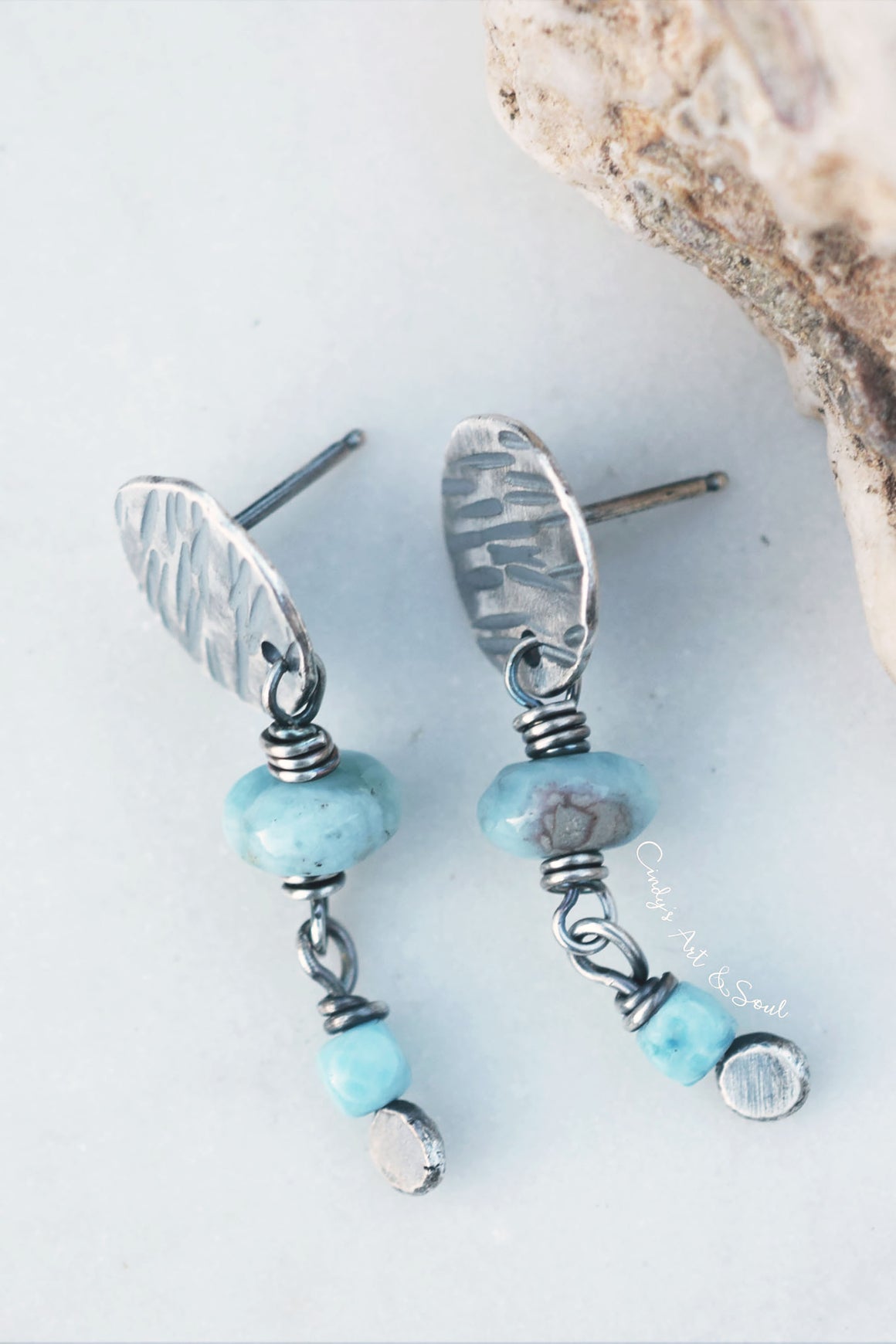 A pair of handmade dangle earrings featuring hammered silver-toned metal discs, wrapped wire, light blue beads, and small polished blue and silver beads, displayed on a light gray surface with a piece of natural rock in the corner.