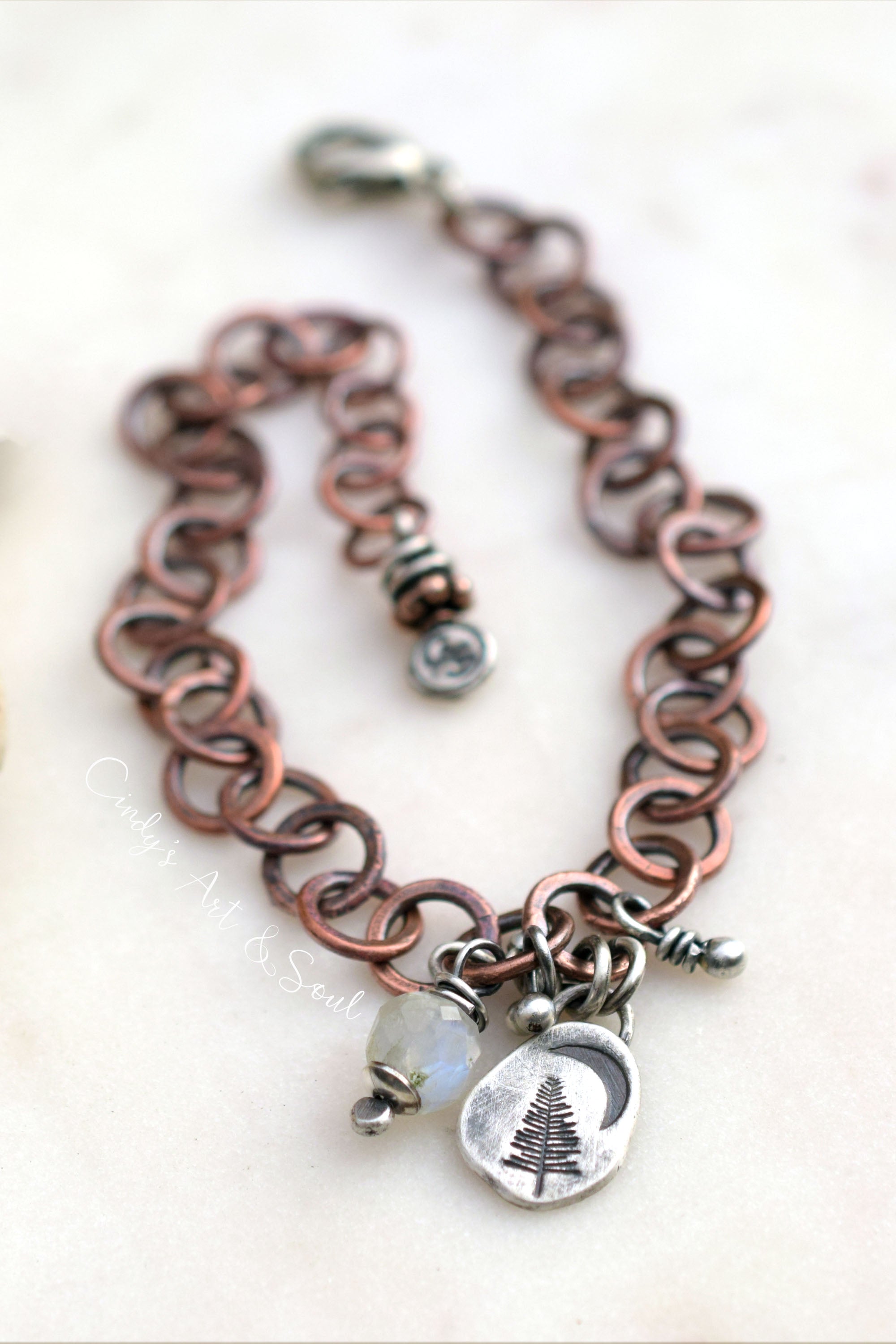 A handmade link chain bracelet in a copper and silver-tone metal, featuring a lobster clasp, a small silver beaded charm, and two dangling charms: a silver crescent moon with a pine tree engraving and a small white opal bead.