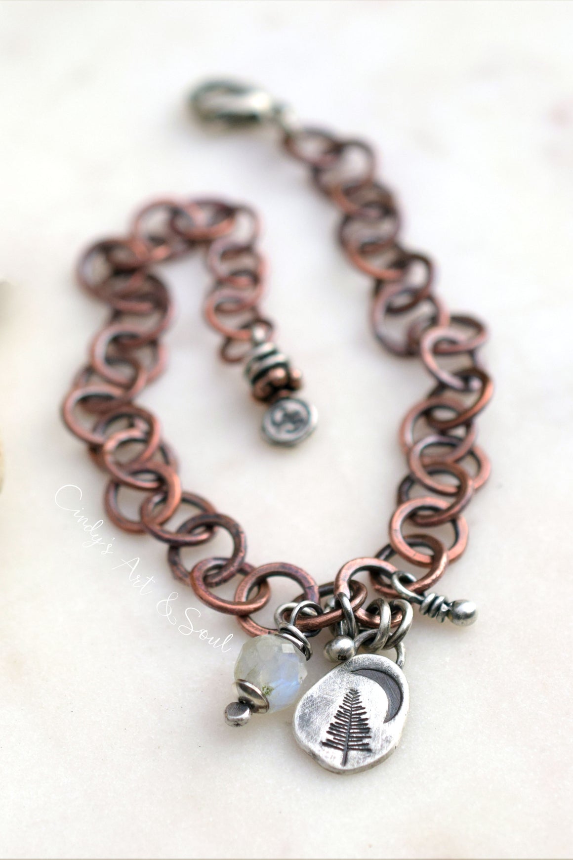 A handmade link chain bracelet in a copper and silver-tone metal, featuring a lobster clasp, a small silver beaded charm, and two dangling charms: a silver crescent moon with a pine tree engraving and a small white opal bead.