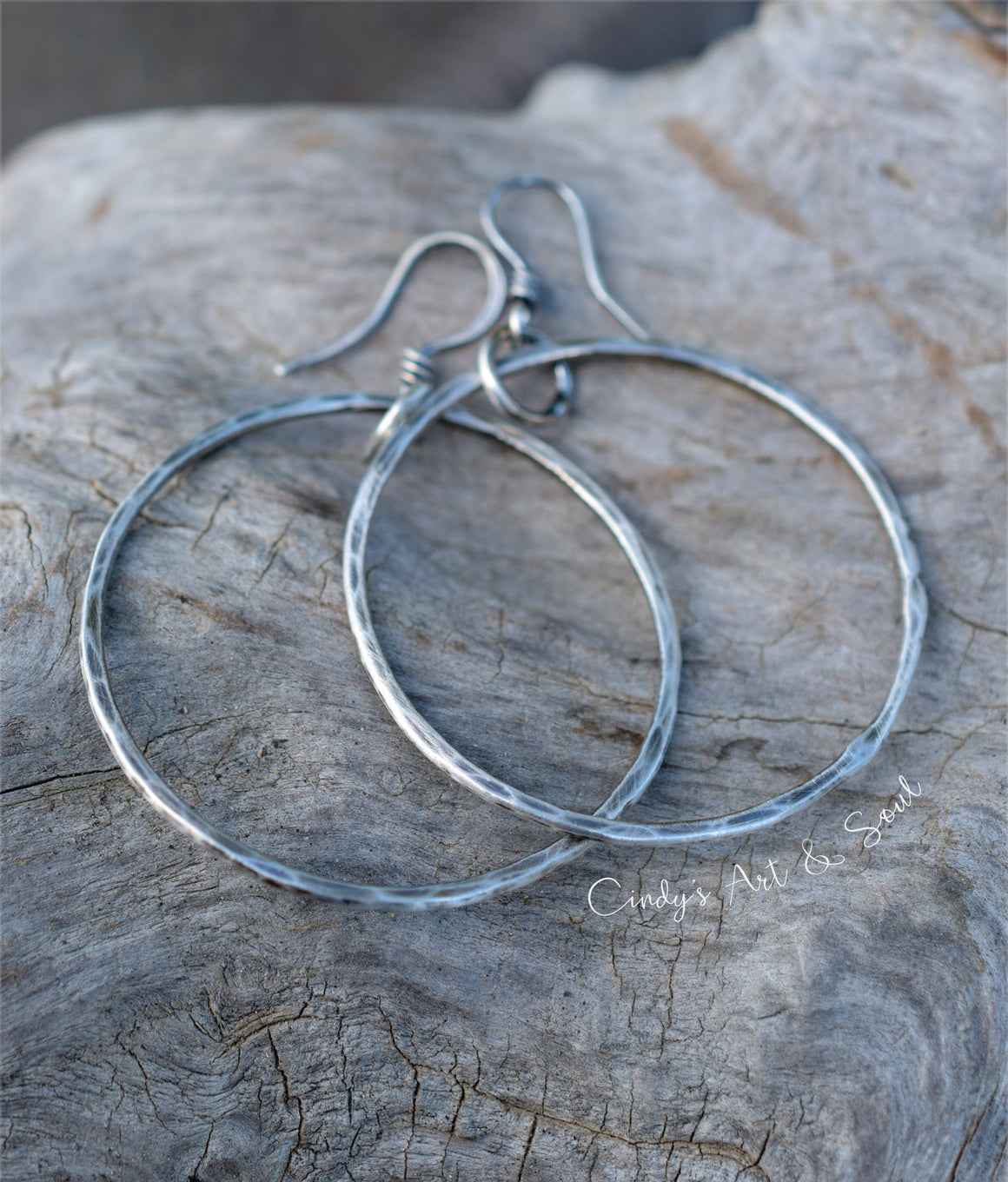 A pair of hammered silver hoop earrings with fishhook backs, displayed on a textured, weathered gray wood surface. A watermark for 'Cindy's Art & Soul' is visible on the lower right.