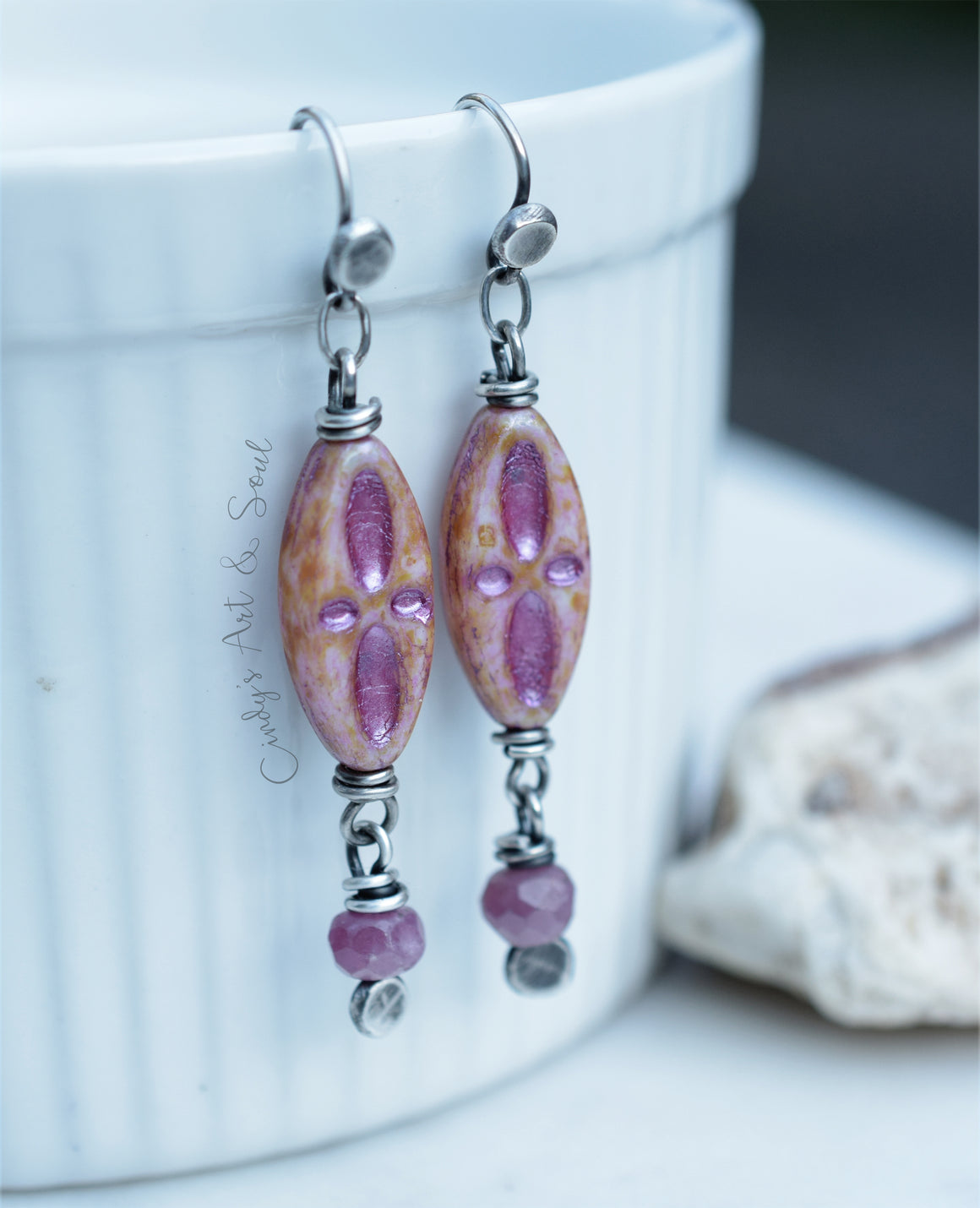 A pair of handmade dangle earrings with oval pink marbled beads and pink ruby faceted beads, hanging from silver fishhook wires against a white ribbed ceramic cup.
