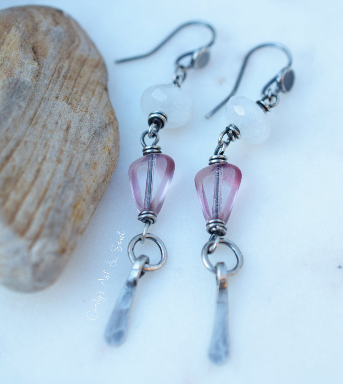 A pair of handmade dangle earrings with faceted pink beads, white round beads, and silver metal hooks and rings, displayed next to a natural stone on a white surface.