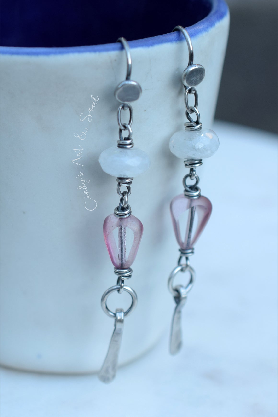 A pair of handmade dangle earrings with silver metal links, a round white faceted bead, a faceted pink bead, and a small silver teardrop charm, hanging from the rim of a white ceramic mug with a blue interior.
