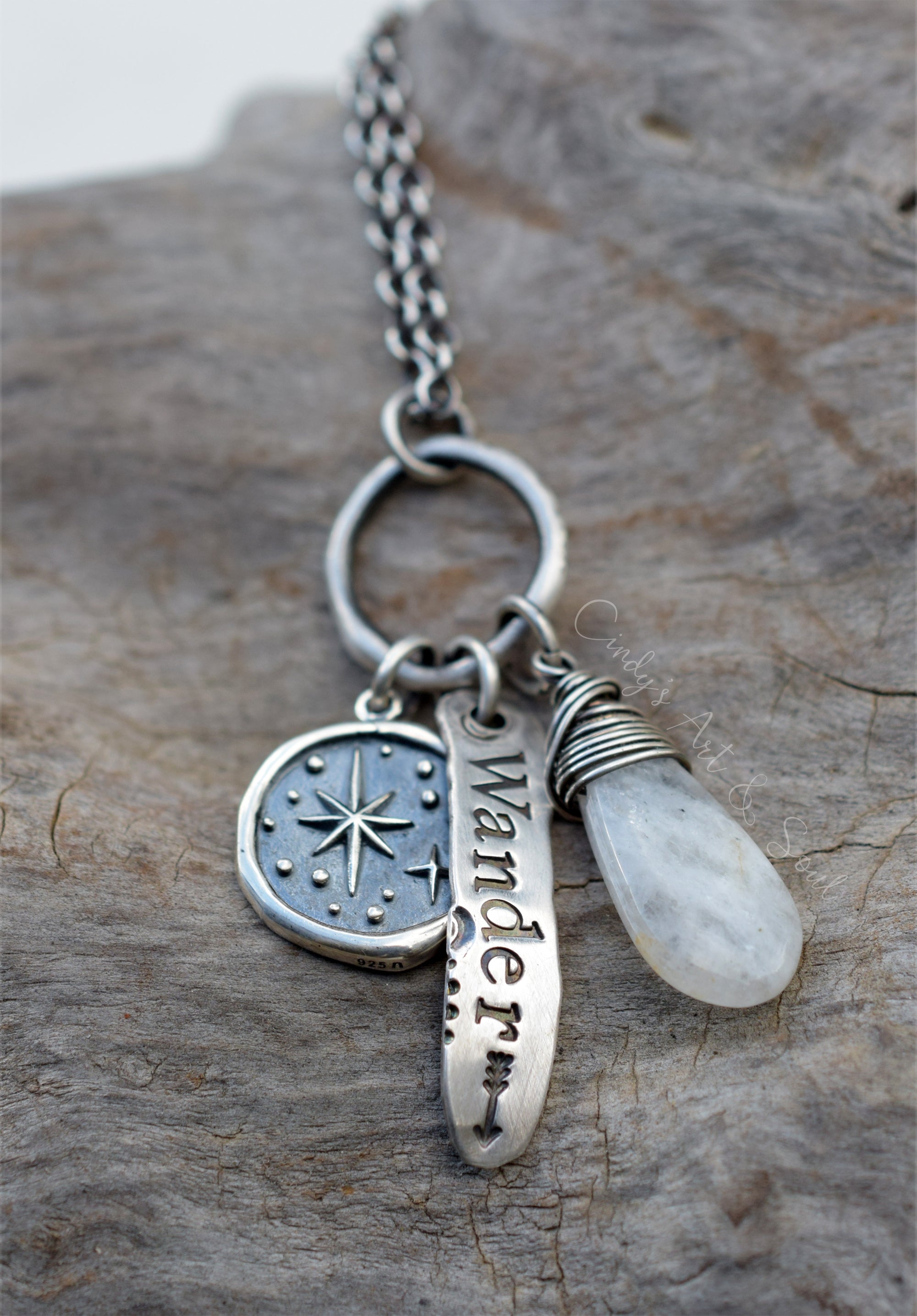 A silver charm necklace with three pendants: a compass, a bar engraved with the word 'Wander', and a teardrop-shaped white moonstone gemstone, displayed on a textured gray stone surface.