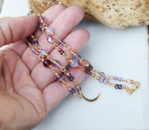 A hand holding crescent moon necklace with golden bronze metal. Beaded chain with beads in various shades of purple.