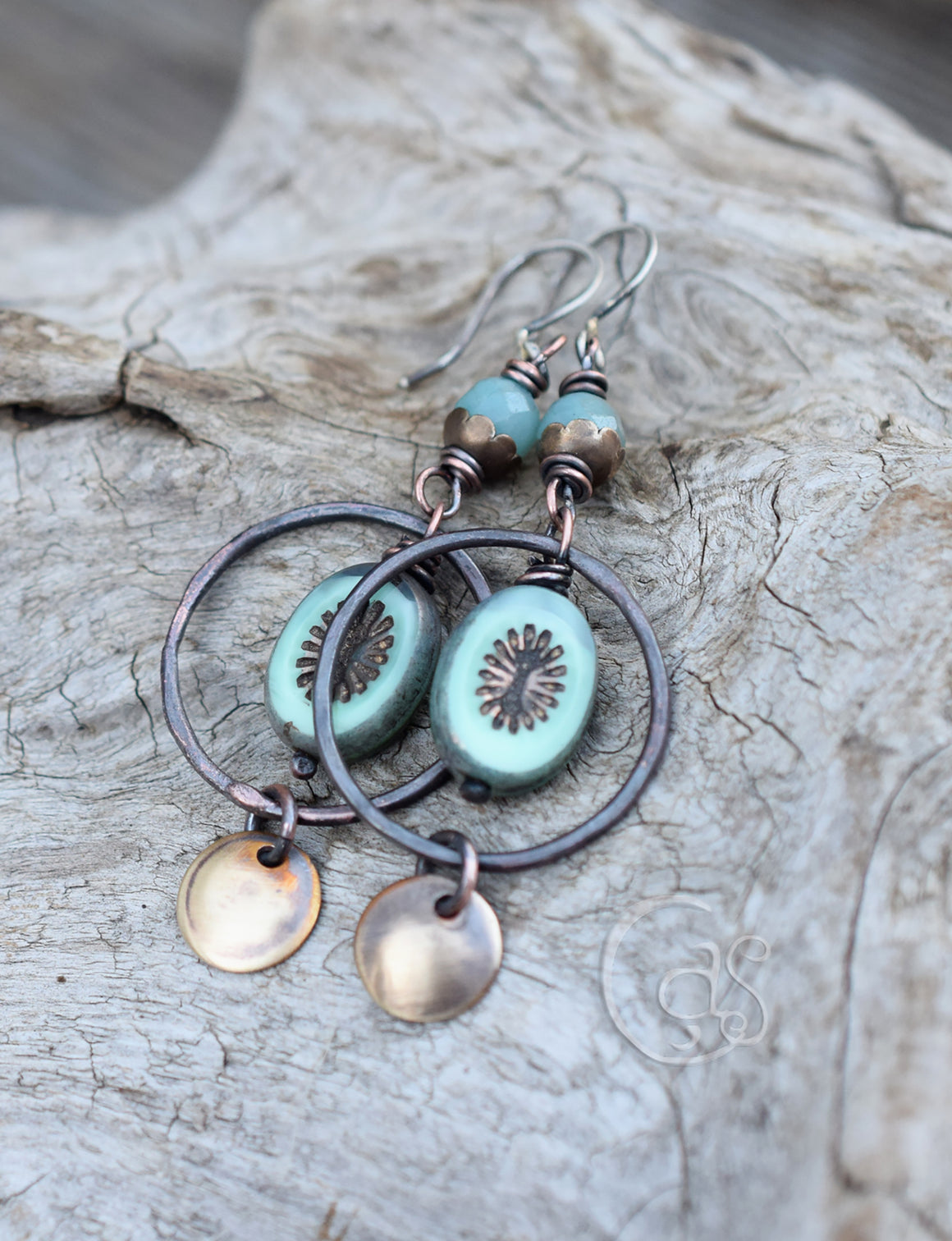 Rusty copper hoop earrings with blue beads that have golden star bursts on them. golden charms dangle below. The top of the earrings have blue stone beads with brass flower caps. The ear wires are silver. These earrings are laying flat on a piece of driftwood. 