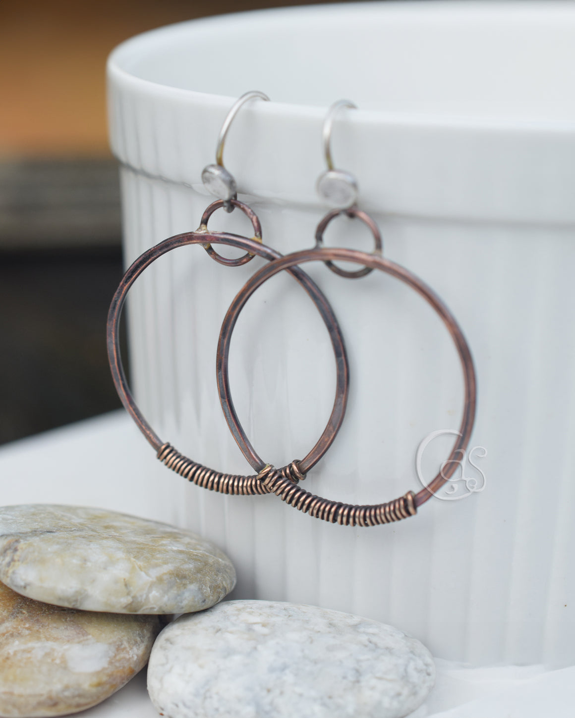 A pair of copper hoop earrings with a hammered, oxidized finish, wrapped in golden bronze wire, displayed on a white surface with a white ceramic cup and some rocks.