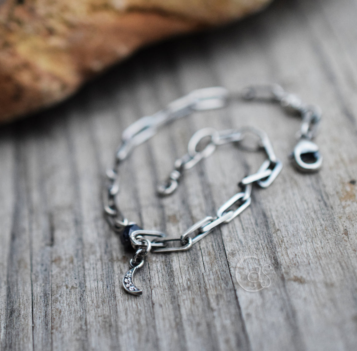 Silver bracelet with blue gemstone and crescent moon charm on a wood background next to a brown rock. 