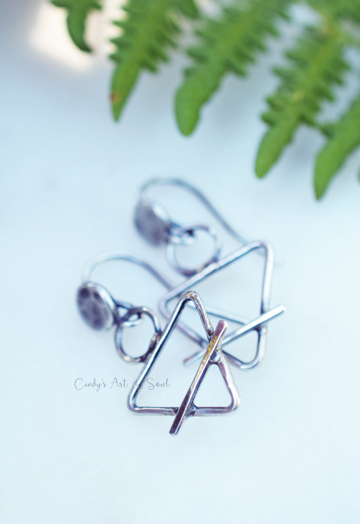 Triangle, earth element silver earrings on a solid white background a fern leaf at the top. 