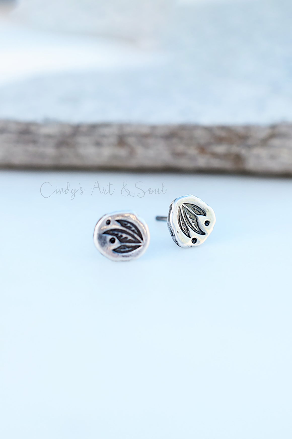 A pair of round silver-toned stud earrings with black oxidized leaf-like detailing, displayed on a light blue surface with a blurred branch in the background. A watermark reading 'Cindy's Art & Soul' is visible across the center.