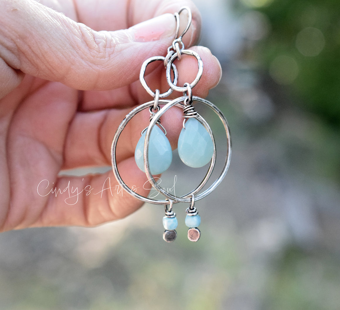 Silver hoop earrings with blue teardrop beads held by a hand against a blurred natural background.