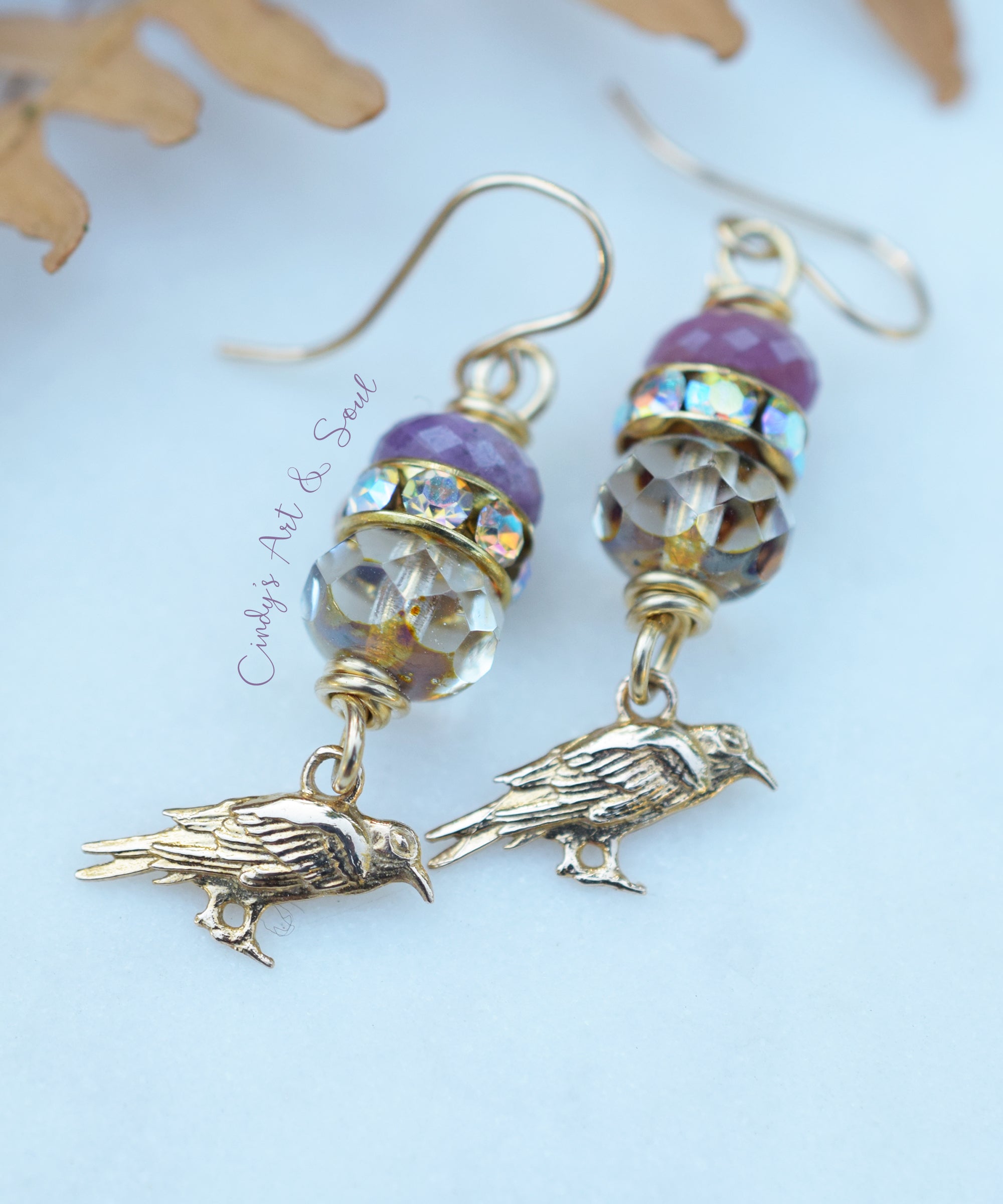 A pair of handmade dangle earrings featuring gold-toned metal hooks and bird charms, embellished with purple beads, faceted clear beads, and iridescent accents, displayed on a light blue background with dried leaves.
