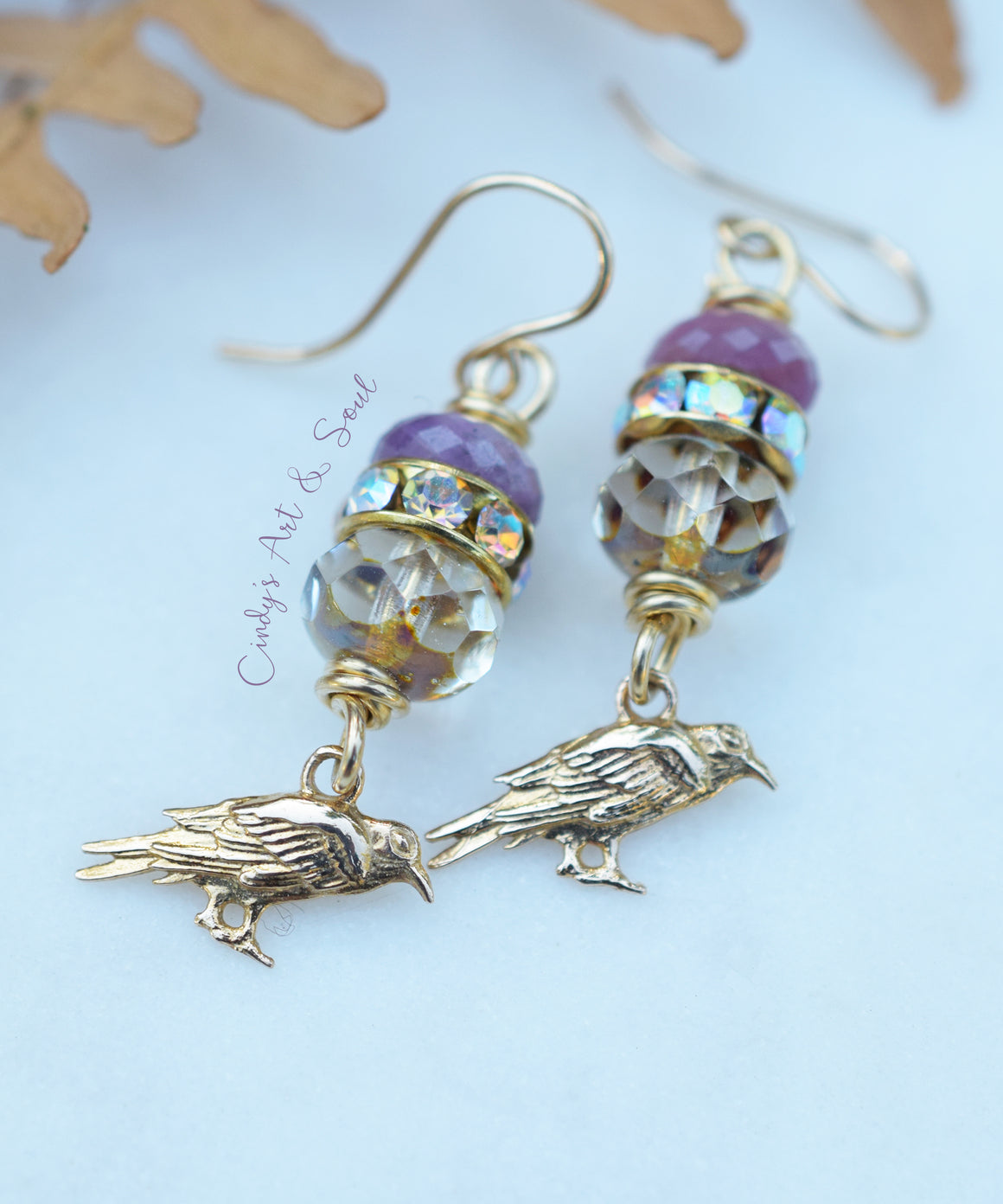 A pair of handmade dangle earrings featuring gold-toned metal hooks and bird charms, embellished with purple beads, faceted clear beads, and iridescent accents, displayed on a light blue background with dried leaves.