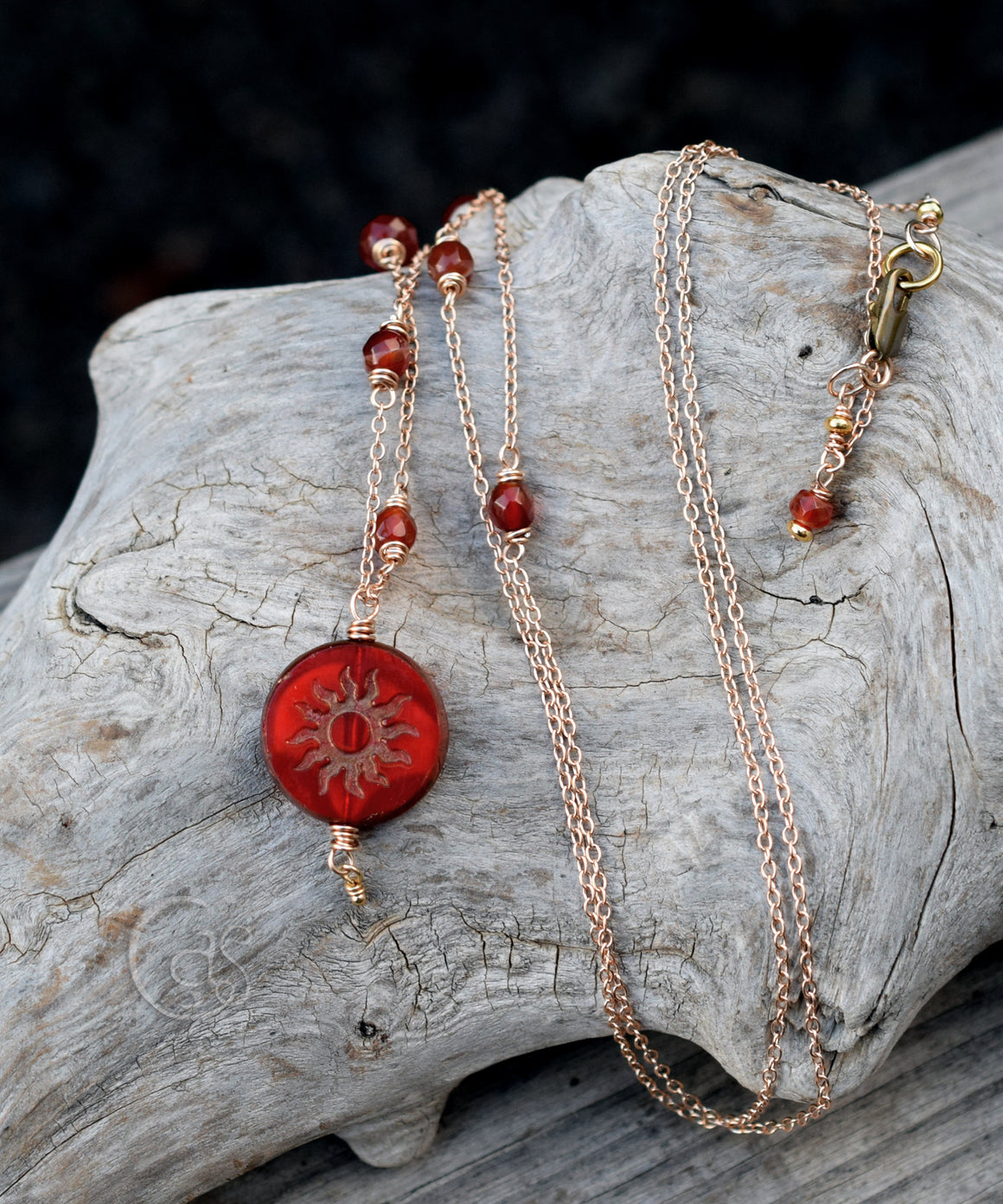 Red gemstones beaded gold chain necklace with a red and gold sun pendant on a piece of driftwood. 
