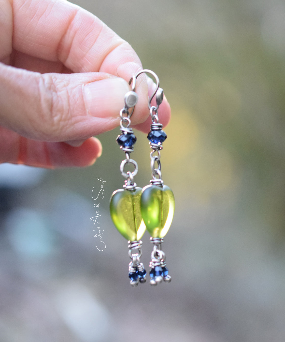 A pair of silver earrings featuring green pressed-glass hearts and dark indigo Swarovski crystals, held between fingers.