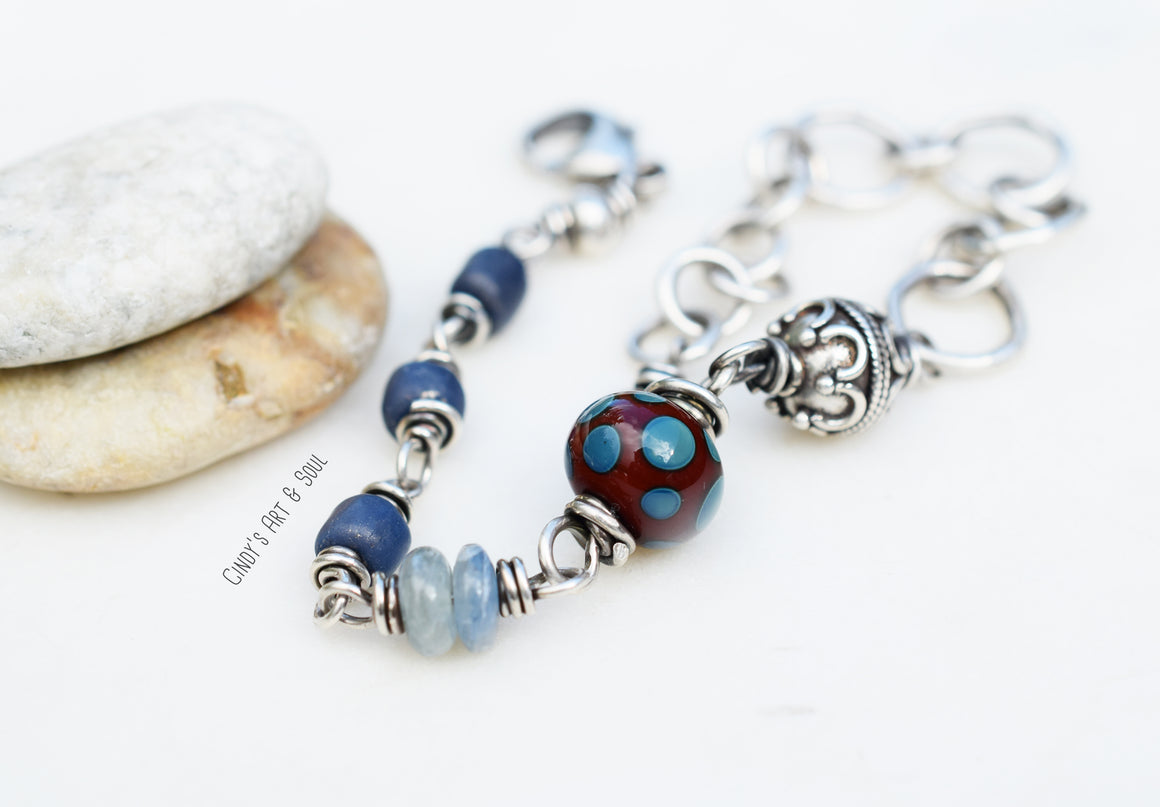 Handcrafted bracelet with a mix of blue and silver beads, connected by intricate silver wire links and featuring a lobster clasp closure. The bracelet includes textured beads, a large dotted focal bead, and delicate chain links, set against a minimalist background with smooth stones.