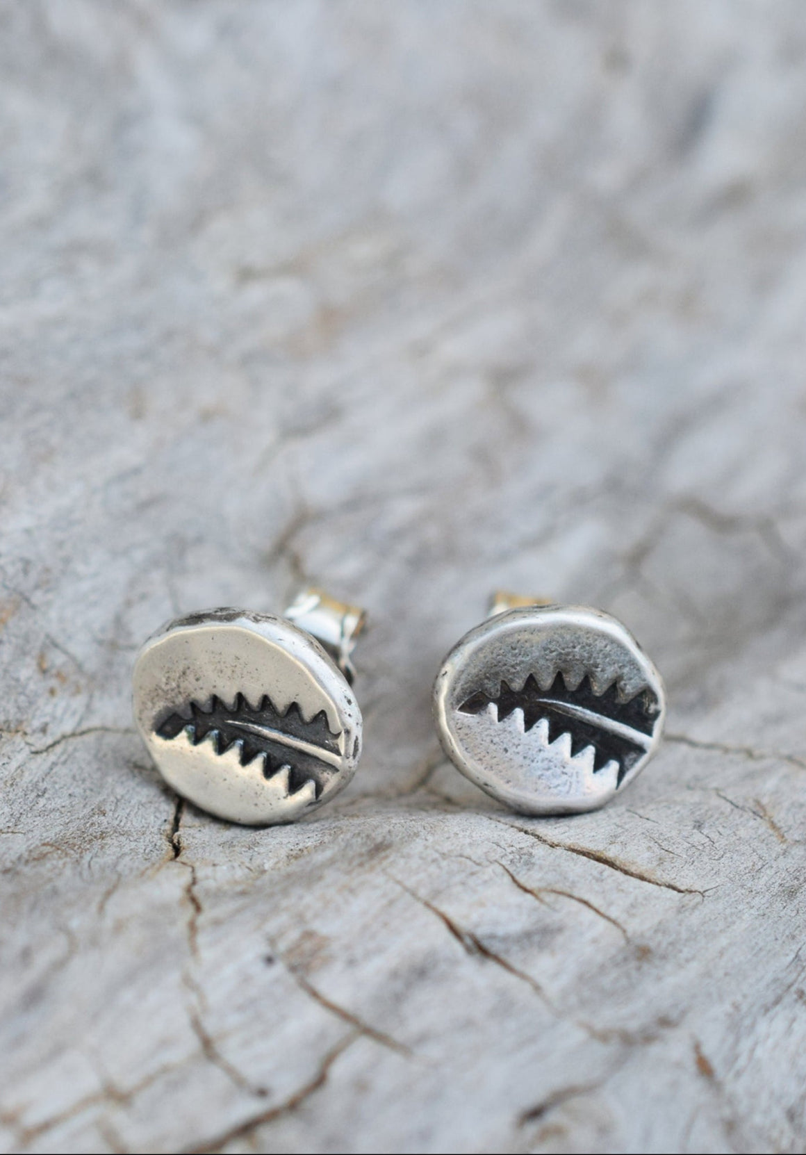 A pair of round silver earrings with a shark-tooth design, resting on a textured, light gray weathered wood surface.