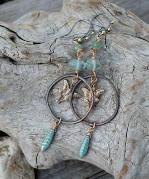 Blackened copper hoops with gold luna moth charms and green gemstones on a wood background. 