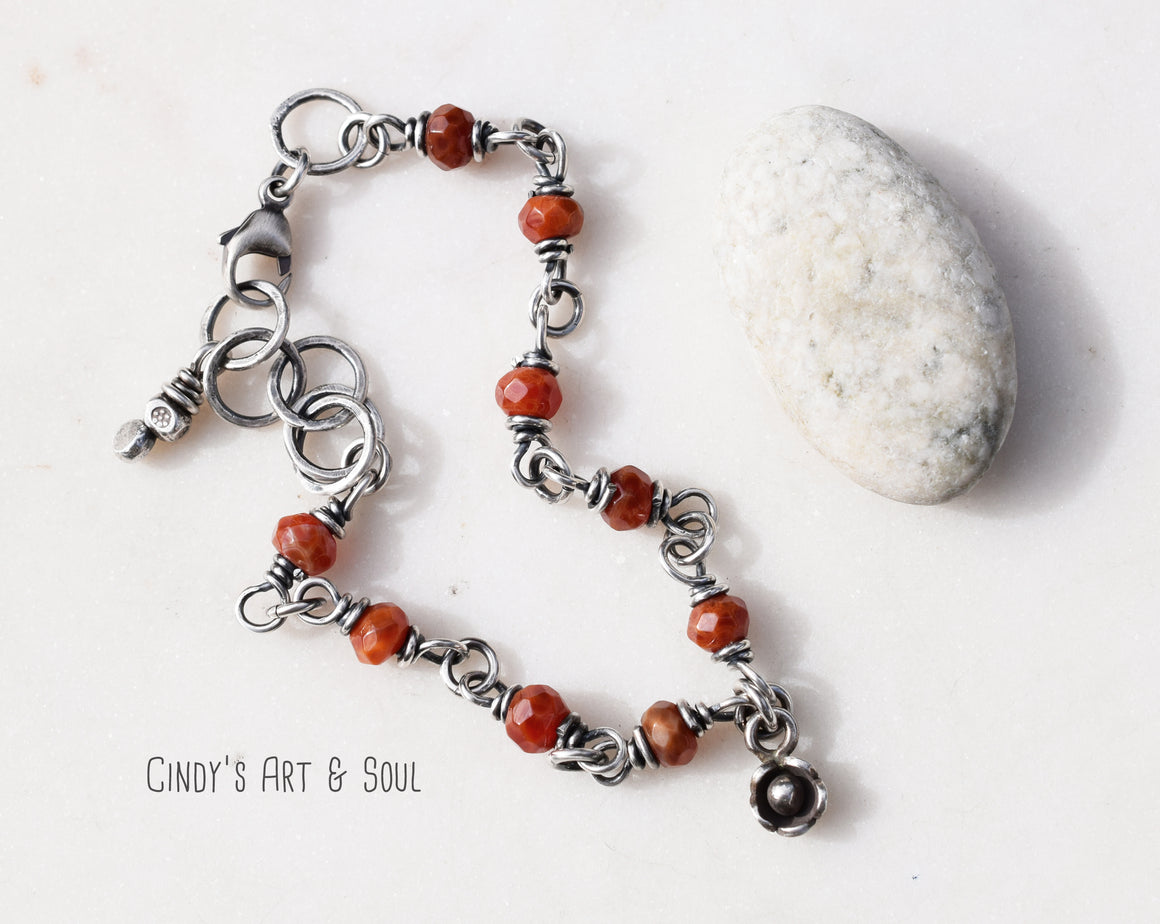 A silver bracelet with dark orange stones next to a white stone on a plain white background. 