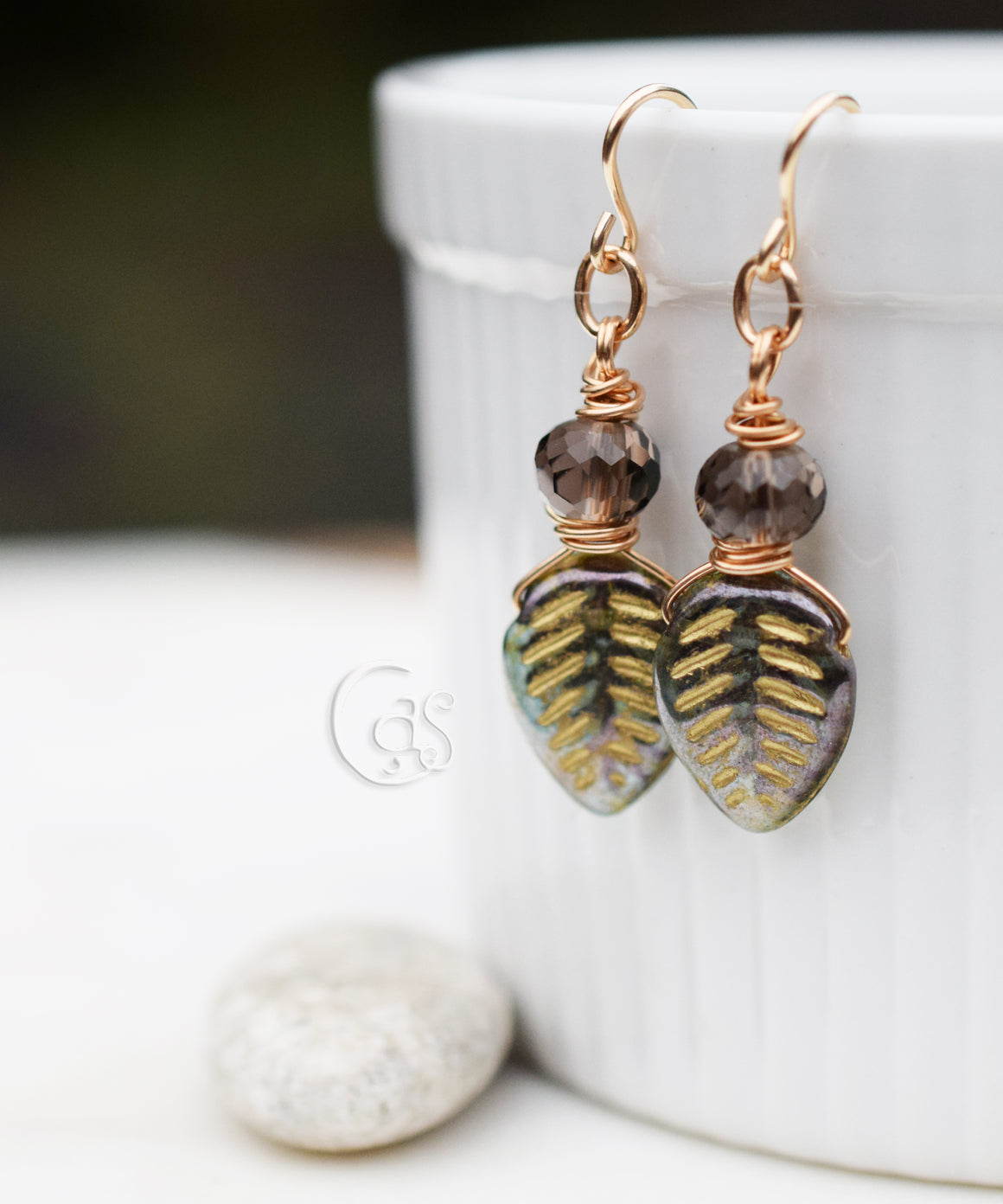 Gold earrings with brown faceted beads and green and gold leaf drops, hanging from a white bowl with a small white stone at the base. 