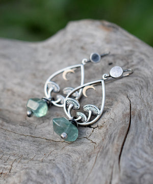 Silver hoop earrings with three mushrooms and a crescent moon in bronze. Green-teal gemstones at the bottom. They sit on a piece of wood with a dark background. 