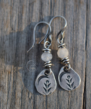 Silver earrings with faceted moonstones and silver charms with a crescent moon and tree stamp. The earrings are on a wood background. 