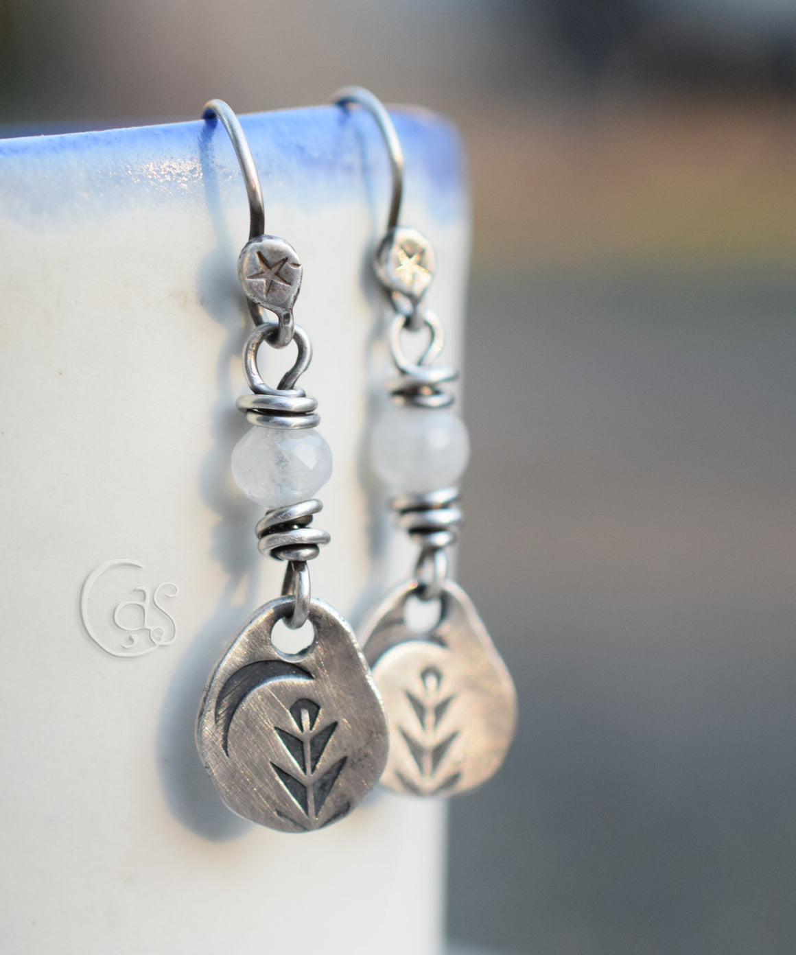Moonstone and silver charm earrings hanging on a white cup. 