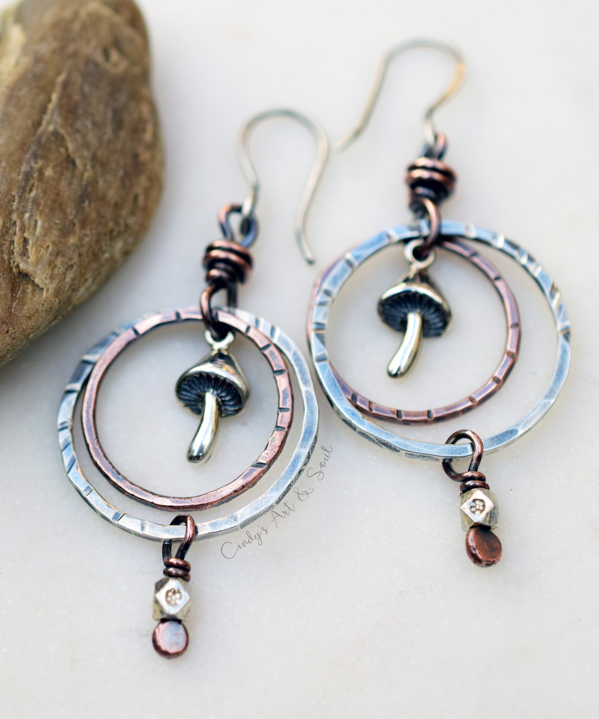 A pair of handmade dangle earrings with hammered silver-tone circular hoops, copper-toned wire wraps, small mushroom charms, and dangling amber and crystal beads, displayed on a light surface with a stone prop.