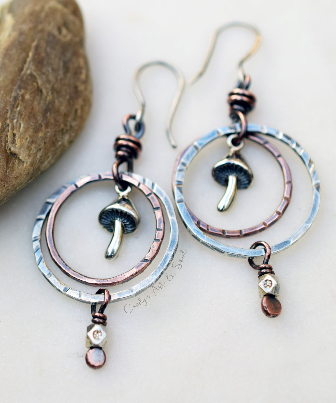 A pair of handmade dangle earrings with hammered silver-tone circular hoops, copper-toned wire wraps, small mushroom charms, and dangling amber and crystal beads, displayed on a light surface with a stone prop.