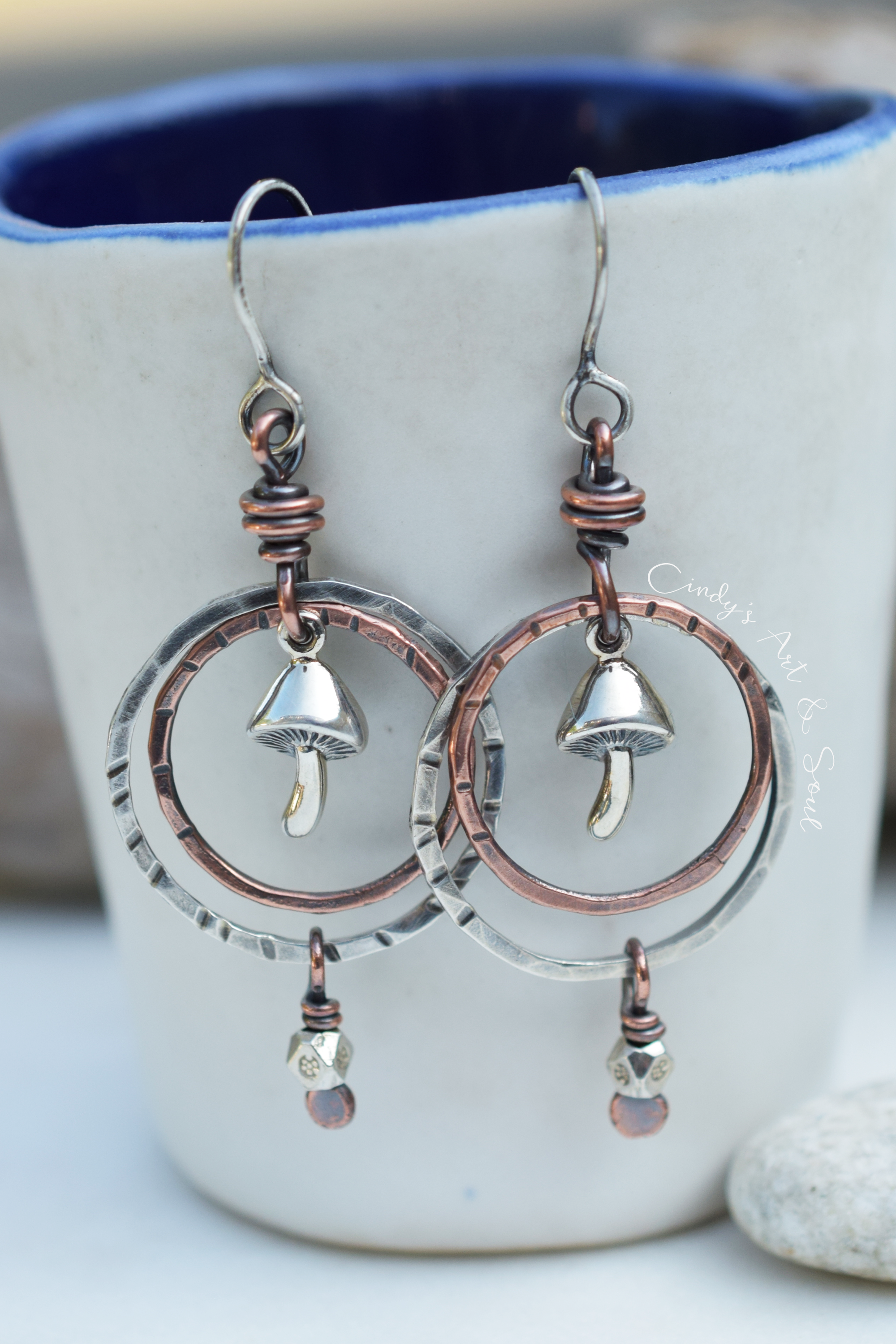 A pair of handmade dangle earrings with hammered silver-tone and copper-tone metal, featuring mushroom charms and circular hoops, hanging in front of a white ceramic mug with a blue interior.