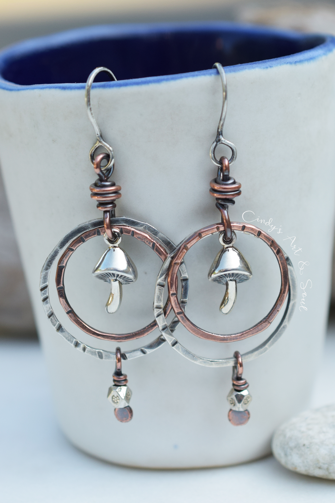 A pair of handmade dangle earrings with hammered silver-tone and copper-tone metal, featuring mushroom charms and circular hoops, hanging in front of a white ceramic mug with a blue interior.