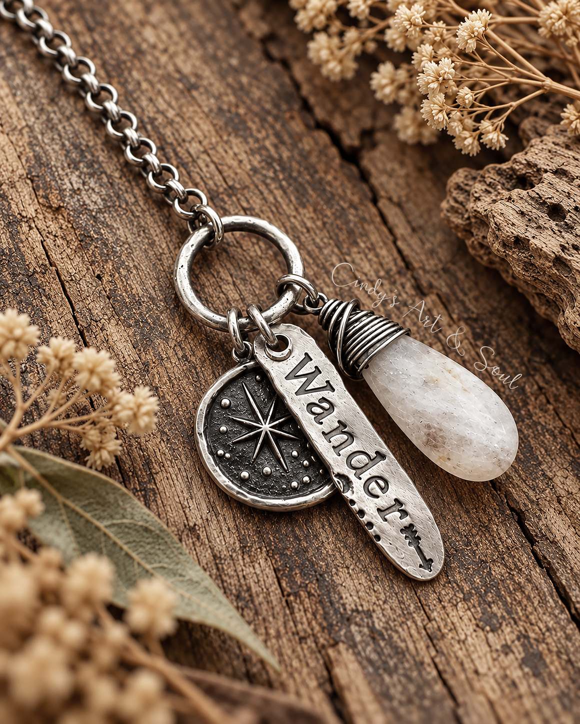 A silver charm necklace with a round ring, a north star compass pendant, a bar engraved with the word 'Wander', and a teardrop-shaped white moonstone gemstone, displayed on a rustic wooden surface with dried flowers.